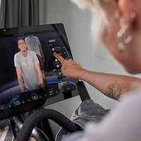 Person using a fitness equipment console with a screen displaying a video of a person exercising.