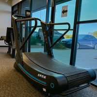 Curved treadmill in a gym with a car visible through large windows.